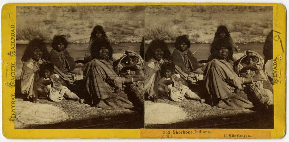 A stereo card showing a group of American Indians from the Shoshone Nation gathered near a river