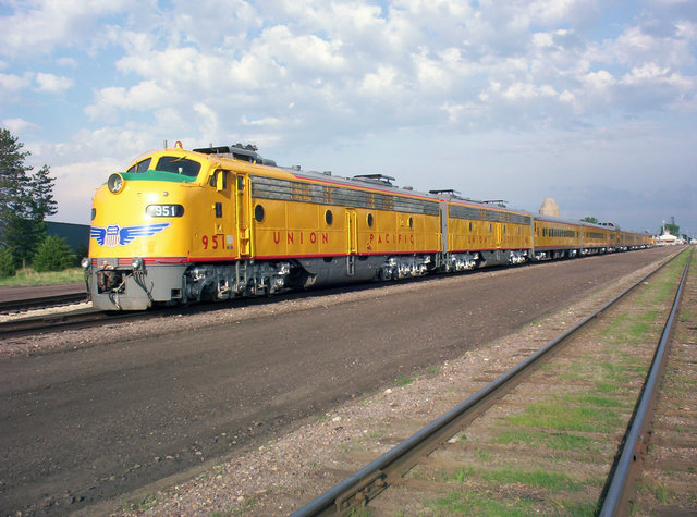 Union Pacific freight train with a yellow locomotive moving through a rural area with grasslands and distant hills, under a blue sky with scattered clouds.
