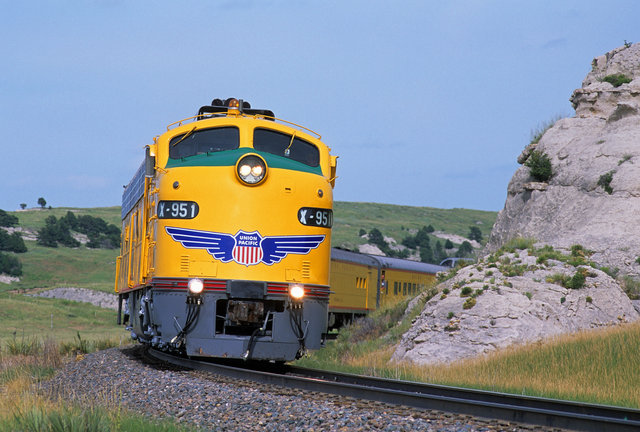 Union Pacific train with multiple yellow locomotives pulling freight cars along railroad tracks in a wide, arid landscape with distant mountains and a partly cloudy sky.