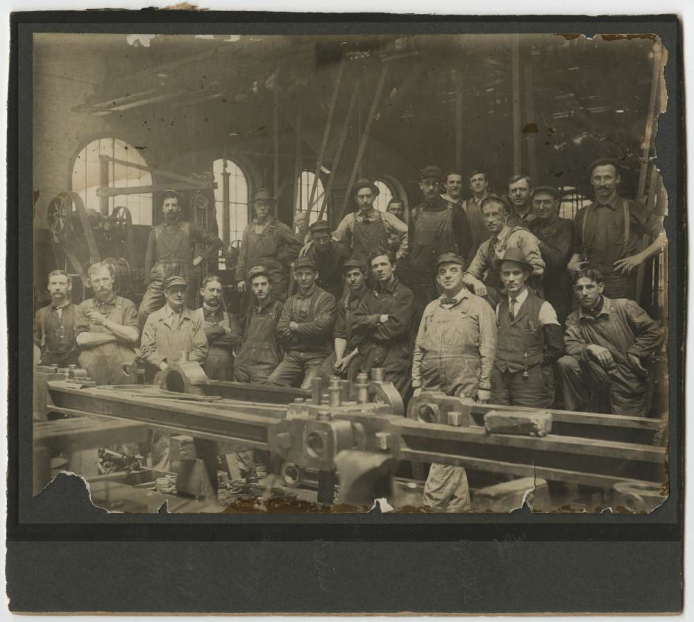 Group of Union Pacific employees pose behind driving rods at locomotive shop in Sacramento, California